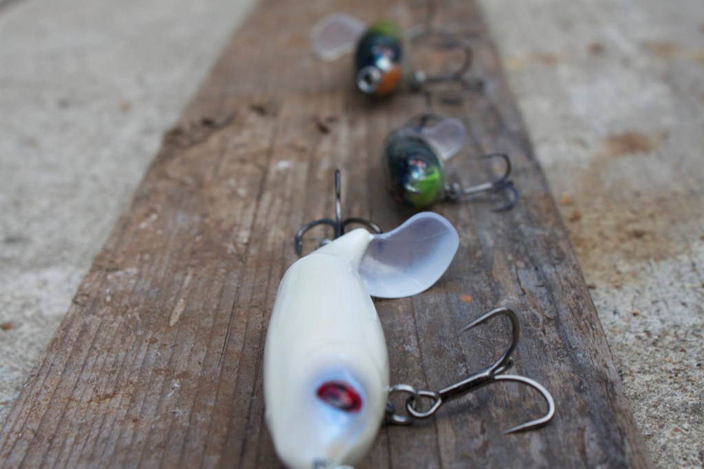 Three compact prop-style bass lures with rotating tail blades arranged on a wooden surface
