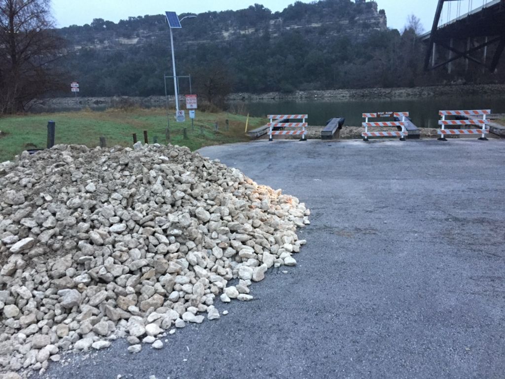 Closed boat ramp with barricades and a pile of rock beside low reservoir water